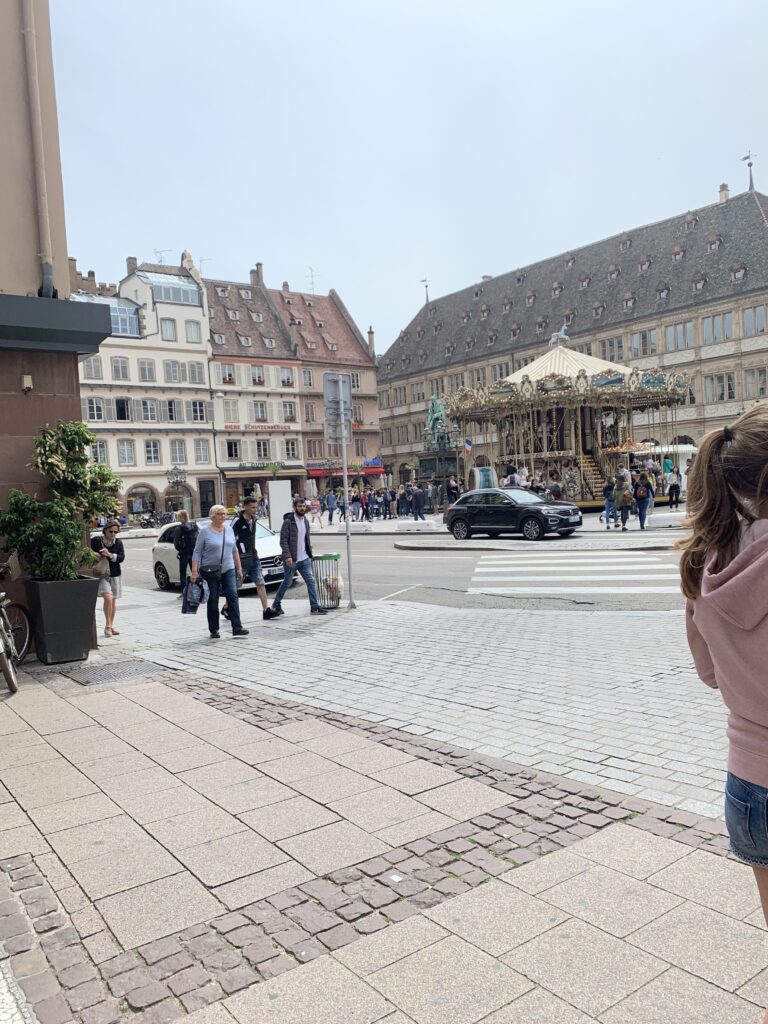 A carousel is located in the center of a large square in Strasbourg, France. Pedestrians and cars move around the carousel.