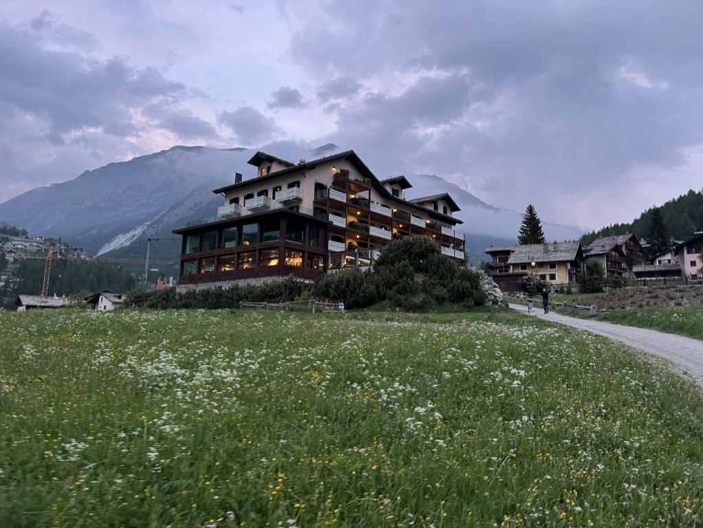 Pink Tyrolean hotel in the center of a mountain region, in Cogne Italy, with a field of white wildflowers in front of it.